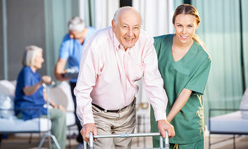 An elderly man using a walker is assisted by a smiling female caregiver in green scrubs, with other elderly individuals engaging in activities in a well-lit indoor setting.