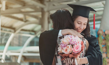 A graduate, in a cap and gown, warmly embraces another person while holding a bouquet of flowers in a modern architectural setting with glass and metal structures.