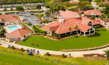 A golf course complex with red-roofed buildings and a swimming pool surrounded by greenery and parked golf carts; cars are visible in a parking lot under sunny skies.