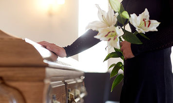 A person holding white lilies places a hand on a wooden coffin in a softly lit room, suggesting a funeral or memorial service.